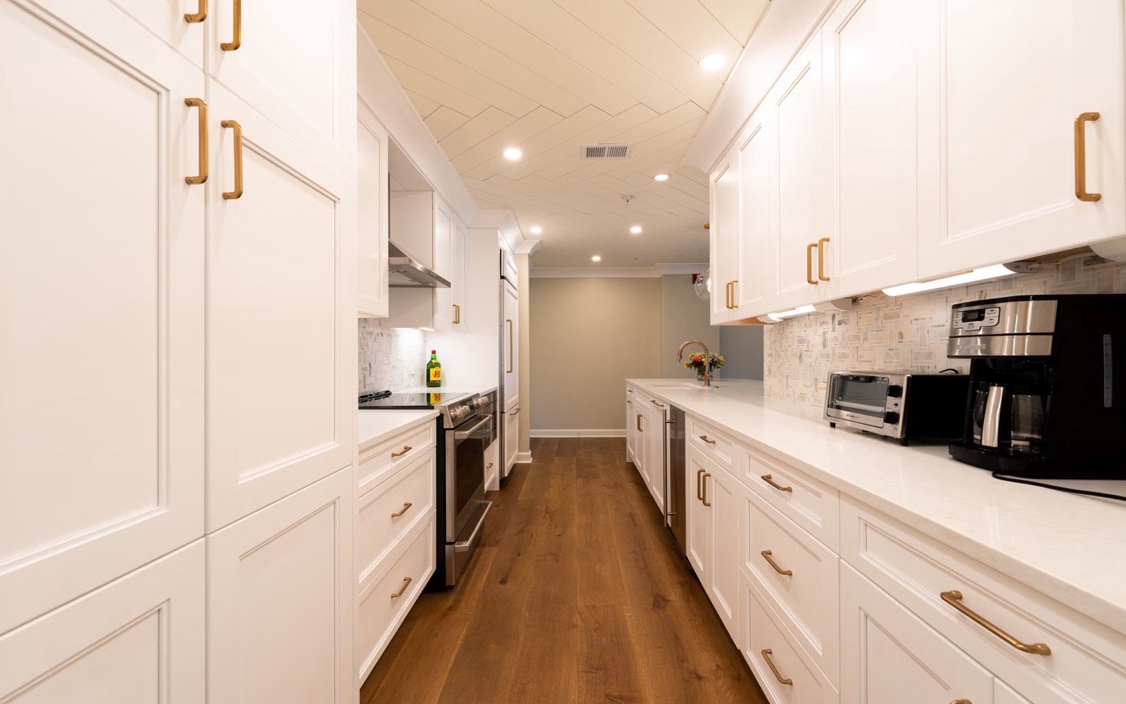 A finished galley kitchen with white shaker cabinets, brass handles and a warm wood floor.